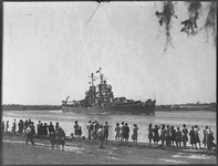 Crowd on shoreline looks on as Navy ship passes by